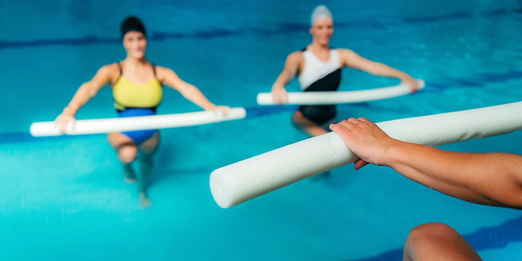 Zwei Erwachsene im Wasser, die mit Schaumstoffstäben trainieren. Eine Person trägt einen gelben und blauen Badeanzug, die andere einen schwarzen Badeanzug mit weißen Akzenten